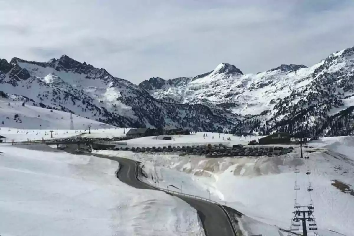 Paisaje montañoso nevado con una carretera que serpentea hacia un estacionamiento lleno de autos y un telesilla en primer plano.