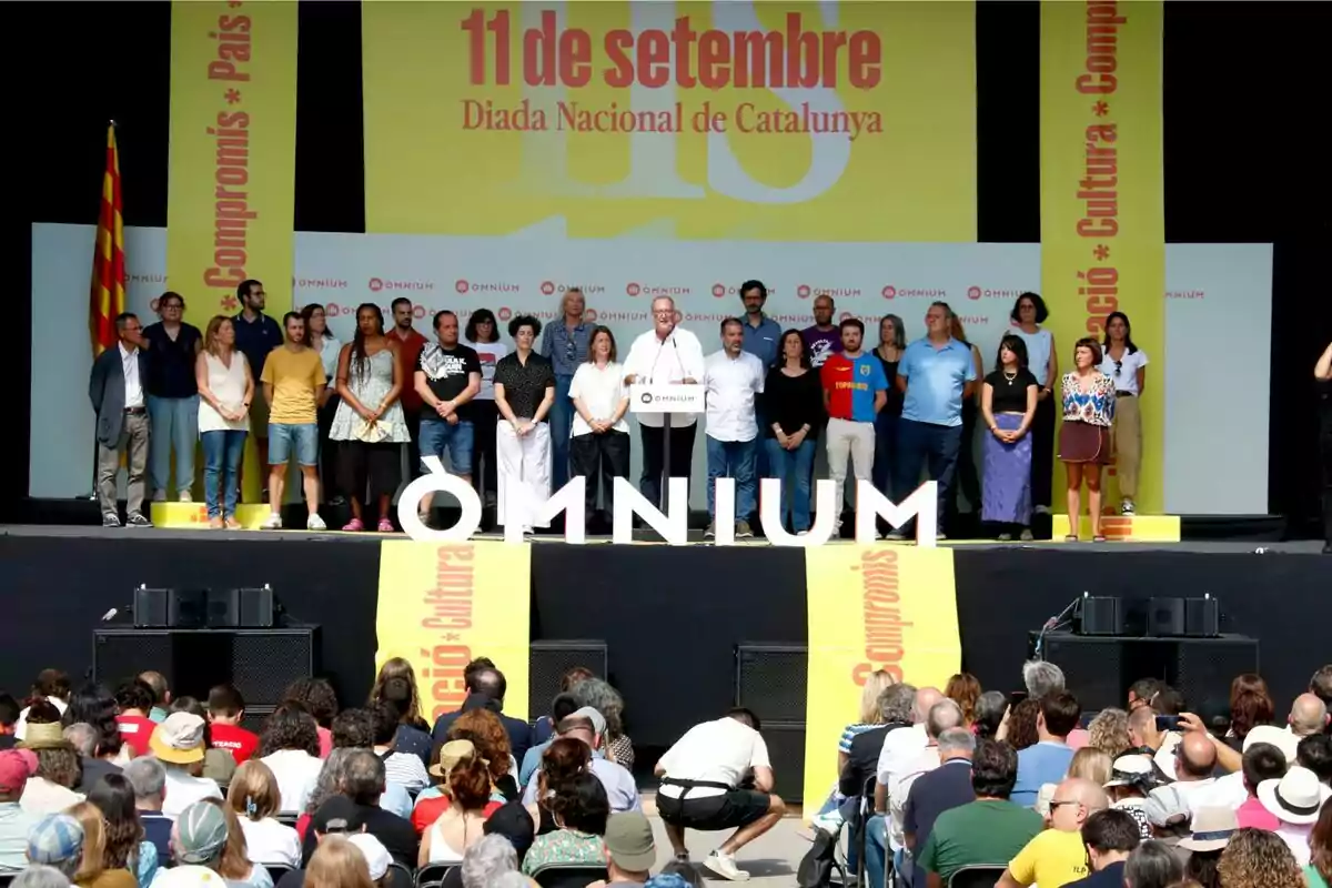 Un grup de persones es troben en un escenari durant la celebració de la Diada Nacional de Catalunya l'11 de setembre, amb una bandera catalana a l'esquerra i pancartes grogues amb el text "Compromís * Cultura * País" als costats.