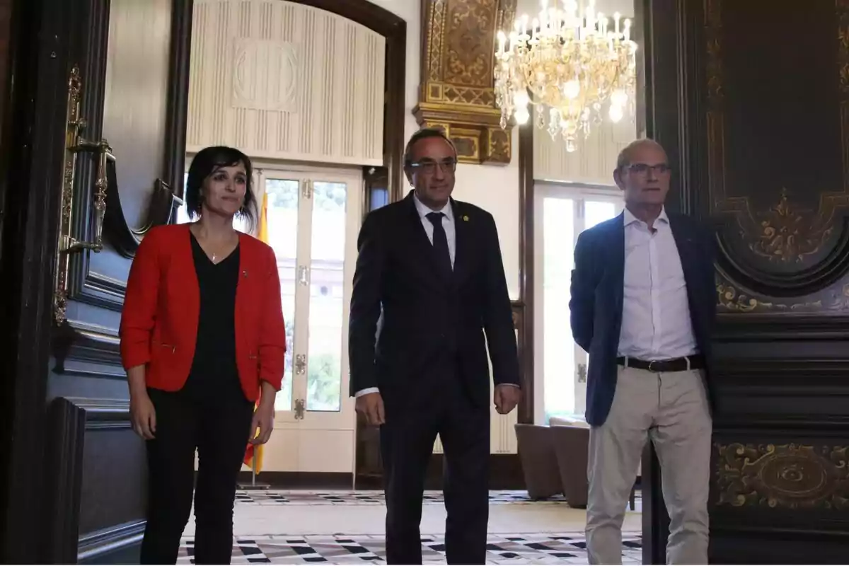 Tres persones posant en una habitació elegant amb un llum d'aranya i detalls decoratius daurats.