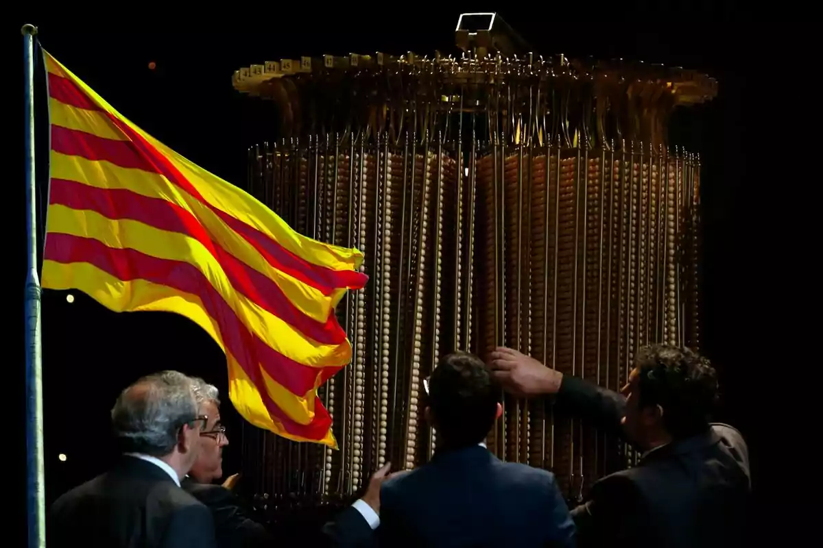 Persones observen una estructura metàl·lica al costat d'una bandera amb franges grogues i vermelles.