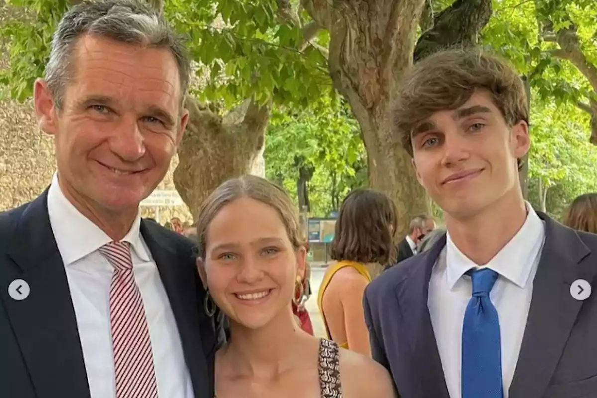 Un hombre de traje y corbata posa junto a una joven y un joven, ambos vestidos formalmente, en un entorno al aire libre con árboles y personas al fondo.