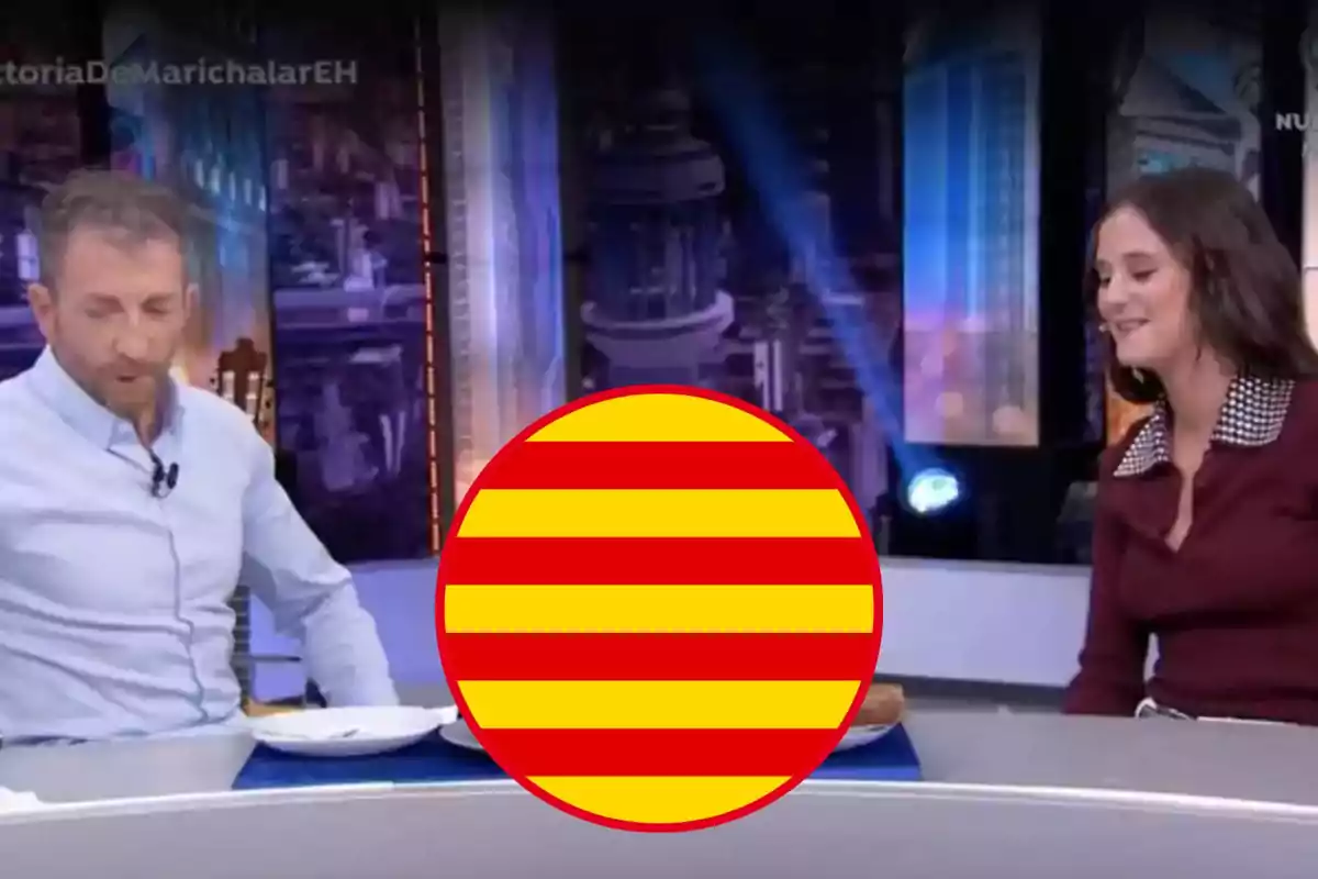 Dos personas están sentadas en una mesa en un programa de televisión, con un círculo con franjas rojas y amarillas superpuesto en el centro de la imagen.