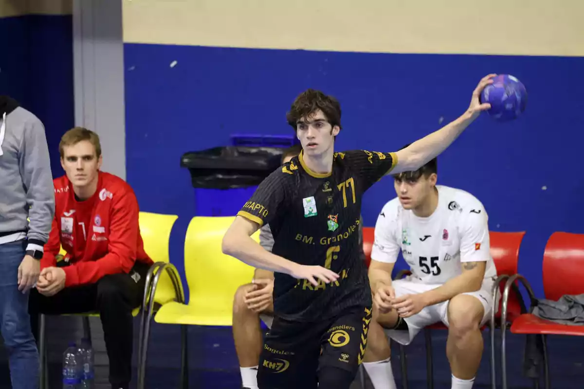 Pablo Urdangarin con uniforme negro se prepara para lanzar el balón mientras otros jugadores están sentados en el banquillo. Pablo Urdangarin con uniforme negro se prepara para lanzar el balón mientras otros jugadores están sentados en el banquillo.
