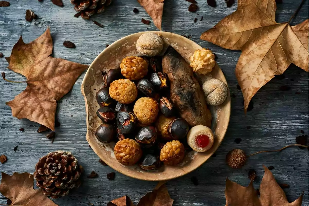 Un plato de castañas asadas, panellets y boniato rodeado de hojas secas y piñas sobre una superficie de madera.