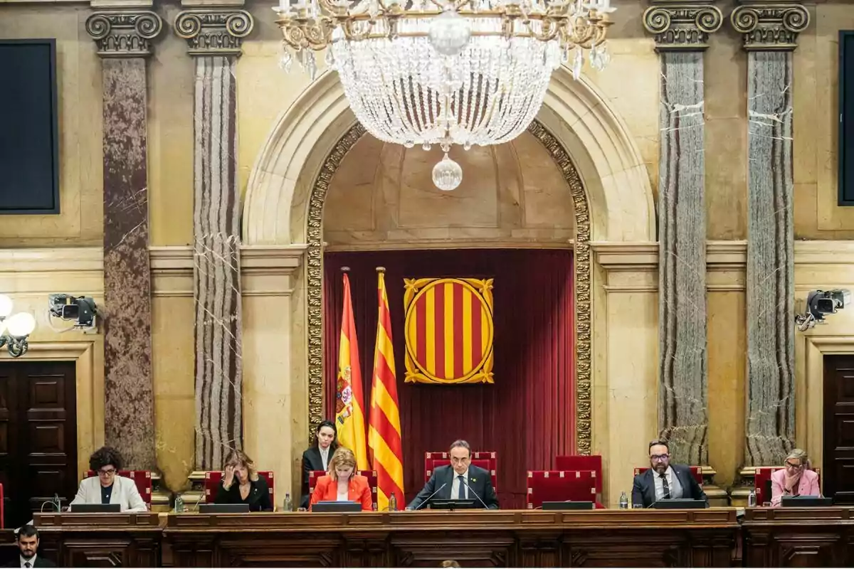 Sessió parlamentària en una sala amb columnes de marbre, una gran làmpada de sostre, banderes d’Espanya i de Catalunya, i diverses persones assegudes a una taula principal. Sessió parlamentària en una sala amb columnes de marbre, una gran làmpada de sostre, banderes d’Espanya i de Catalunya, i diverses persones assegudes a una taula principal.