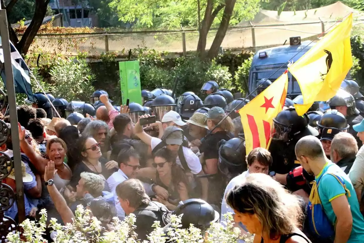 Una multitud de persones s'enfronta a un grup de policies antiavalots, alguns manifestants porten banderes i pancartes mentre els policies estan equipats amb cascos i escuts.