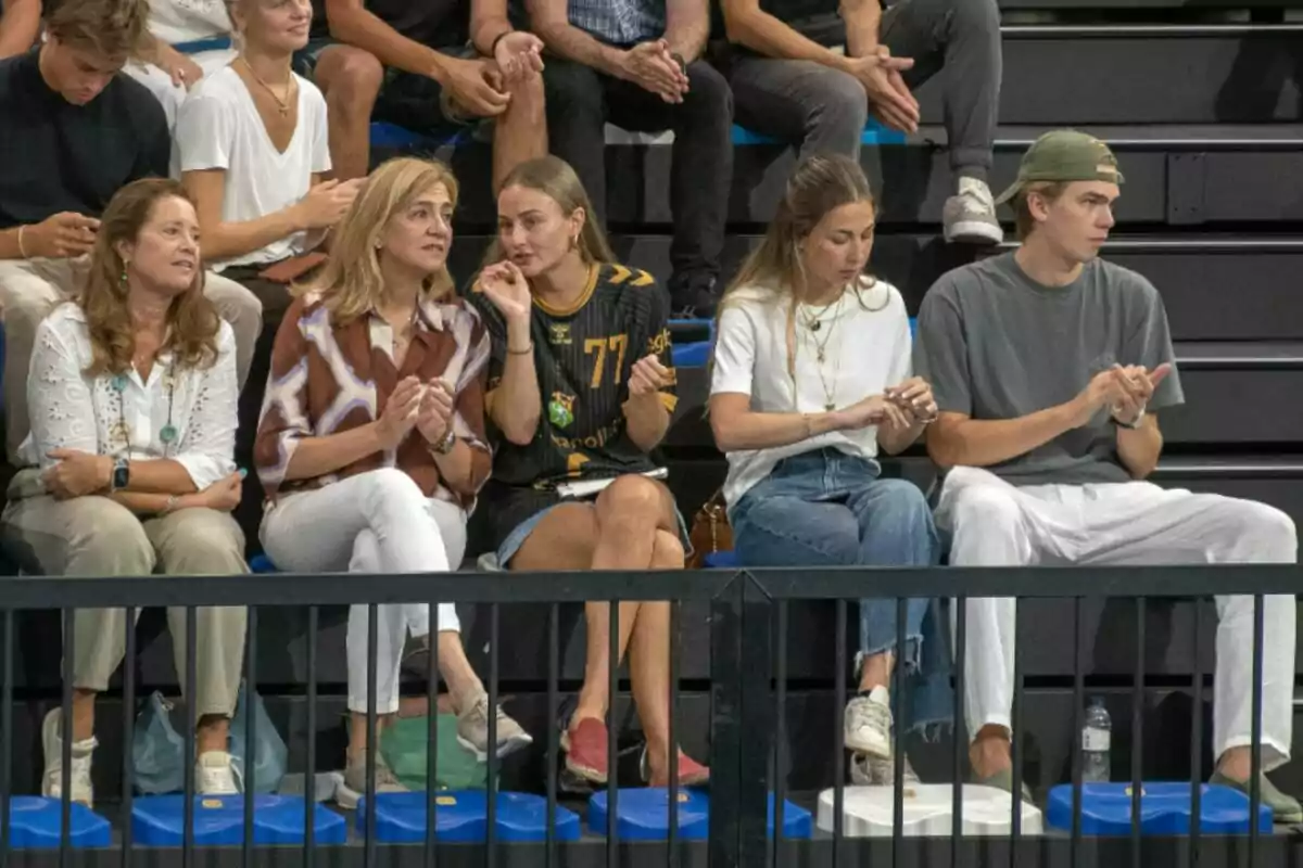 Un grup de persones està assegut a les grades d'un esdeveniment esportiu, algunes aplaudeixen mentre altres semblen conversar o mirar cap al front. Un grup de persones està assegut a les grades d'un esdeveniment esportiu, algunes aplaudeixen mentre altres semblen conversar o mirar cap al front.