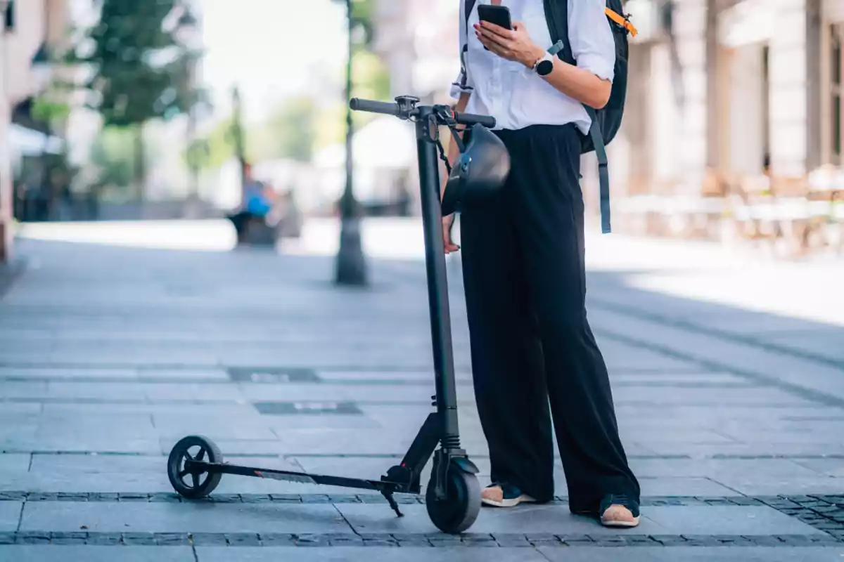 Persona de peu al carrer al costat d'un patinet elèctric mentre revisa el telèfon mòbil.