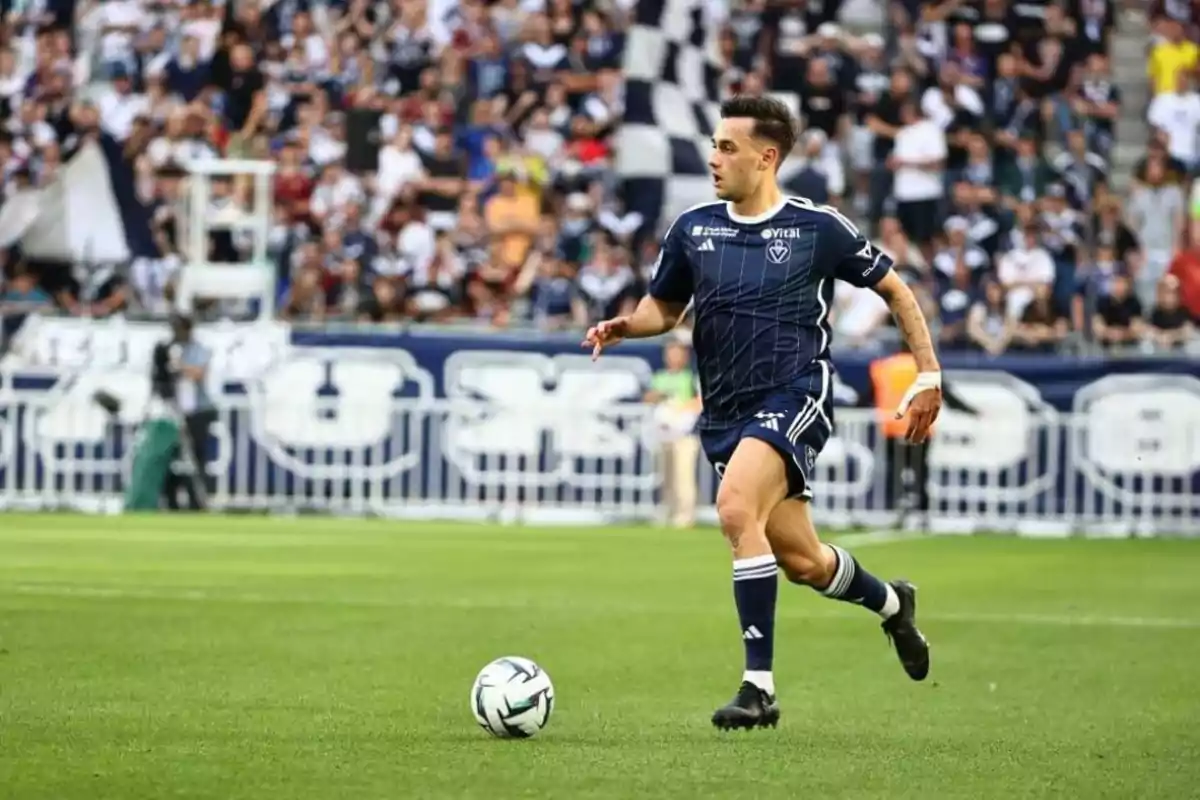 Jugador de futbol en uniforme blau fosc corrent amb la pilota en un estadi ple despectadors.