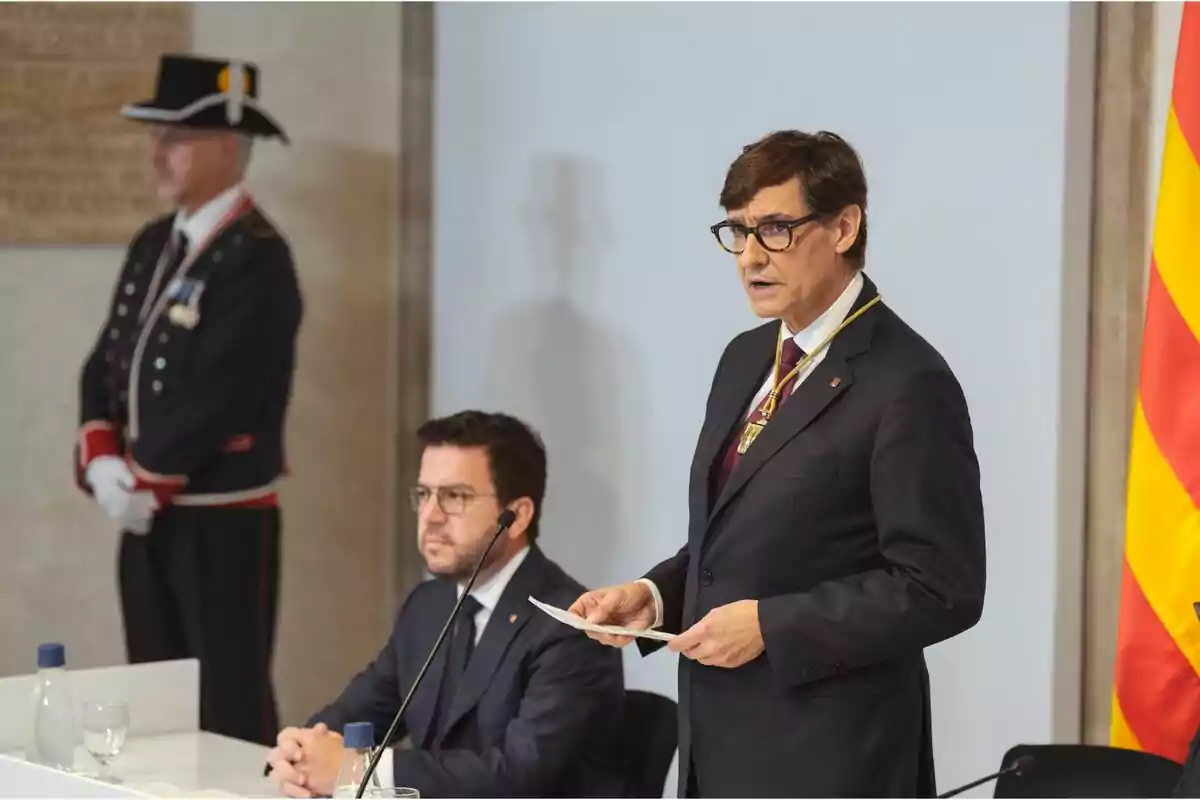 Un home dret amb ulleres i vestit fosc, sostenint un paper, mentre un altre home està assegut al costat amb un micròfon a l'orella; al fons, un guàrdia amb uniforme i una bandera amb franges grogues i vermelles.