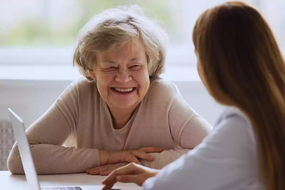 Una mujer mayor sonriendo mientras conversa con otra persona en un entorno interior.