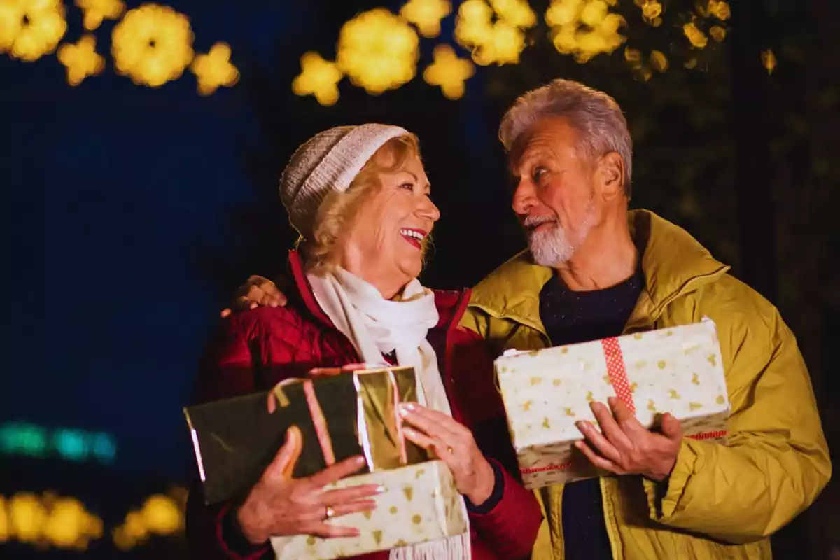 Una pareja mayor sonríe mientras sostiene regalos envueltos, con luces festivas brillando en el fondo.