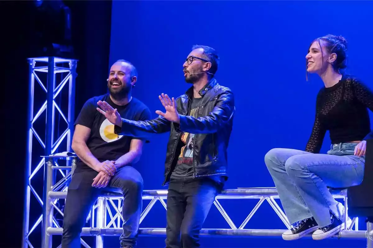 Tres persones estan assegudes en un escenari amb fons blau, dos homes i una dona, tots sembla que estan participant en una conversa animada i divertida.