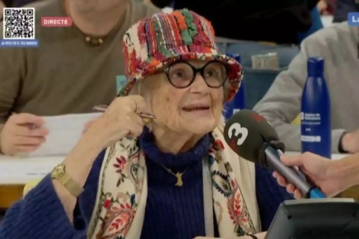 Una persona mayor con sombrero colorido y bufanda estampada es entrevistada por un micrófono en un evento.