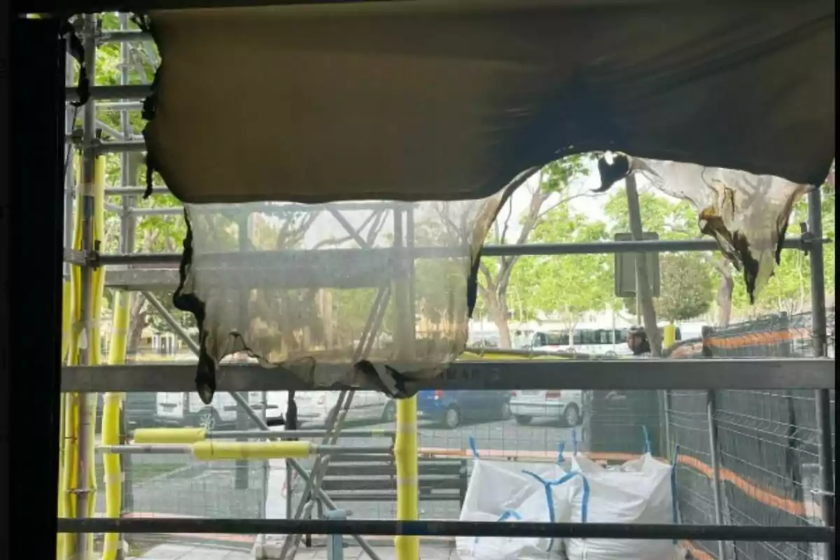 Una lona cremada penja d'una bastida en un lloc de construcció, amb vehicles i arbres visibles al fons.