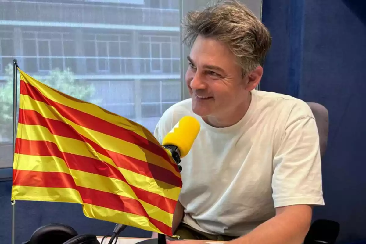 Home amb samarreta blanca parlant en un micròfon groc amb una bandera catalana a primer pla.