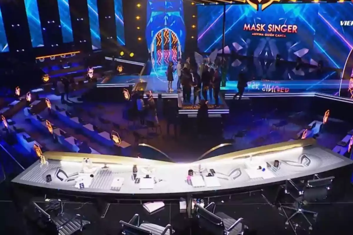 Lit stage of the Mask Singer show with a judges' table in the foreground and several people gathered in the center of the stage Lit stage of the Mask Singer show with a judges' table in the foreground and several people gathered in the center of the stage