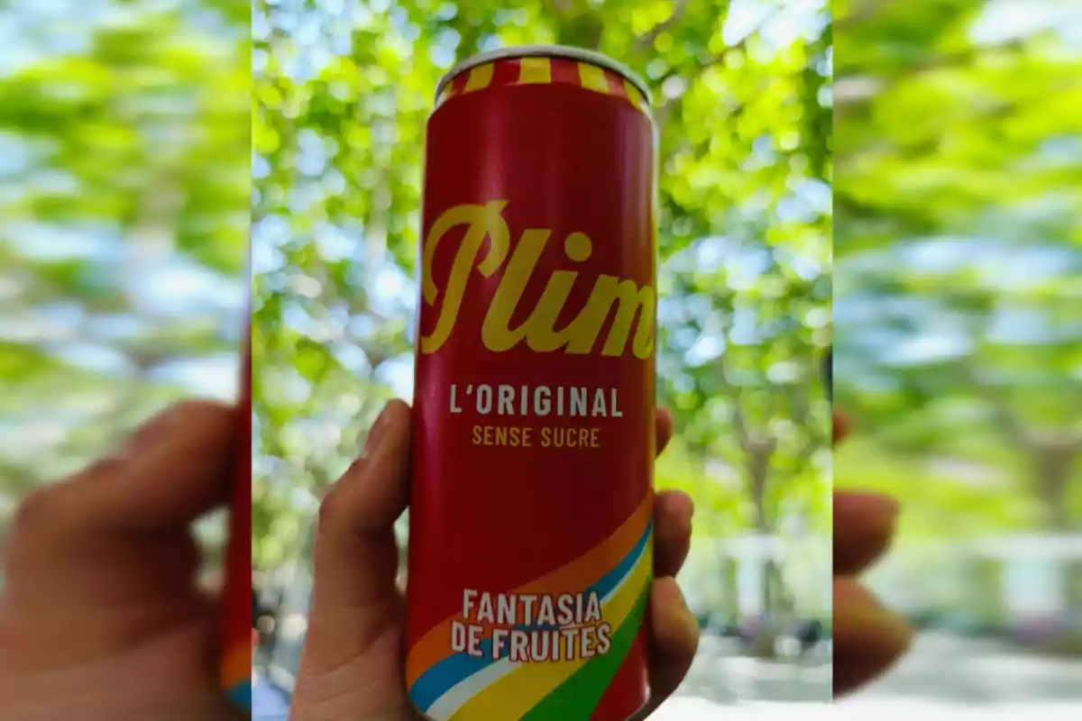 Persona sostenint una llauna vermella de refresc Plim sabor fantasia de fruites amb un fons d’arbres desenfocat Persona sostenint una llauna vermella de refresc Plim sabor fantasia de fruites amb un fons d’arbres desenfocat