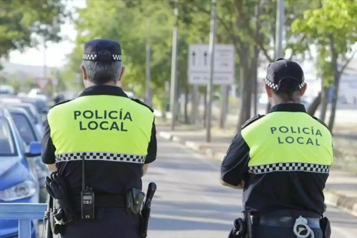 Dos agents de la policia local d'esquena amb armilles reflectores grogues i uniformes negres patrullant un carrer.