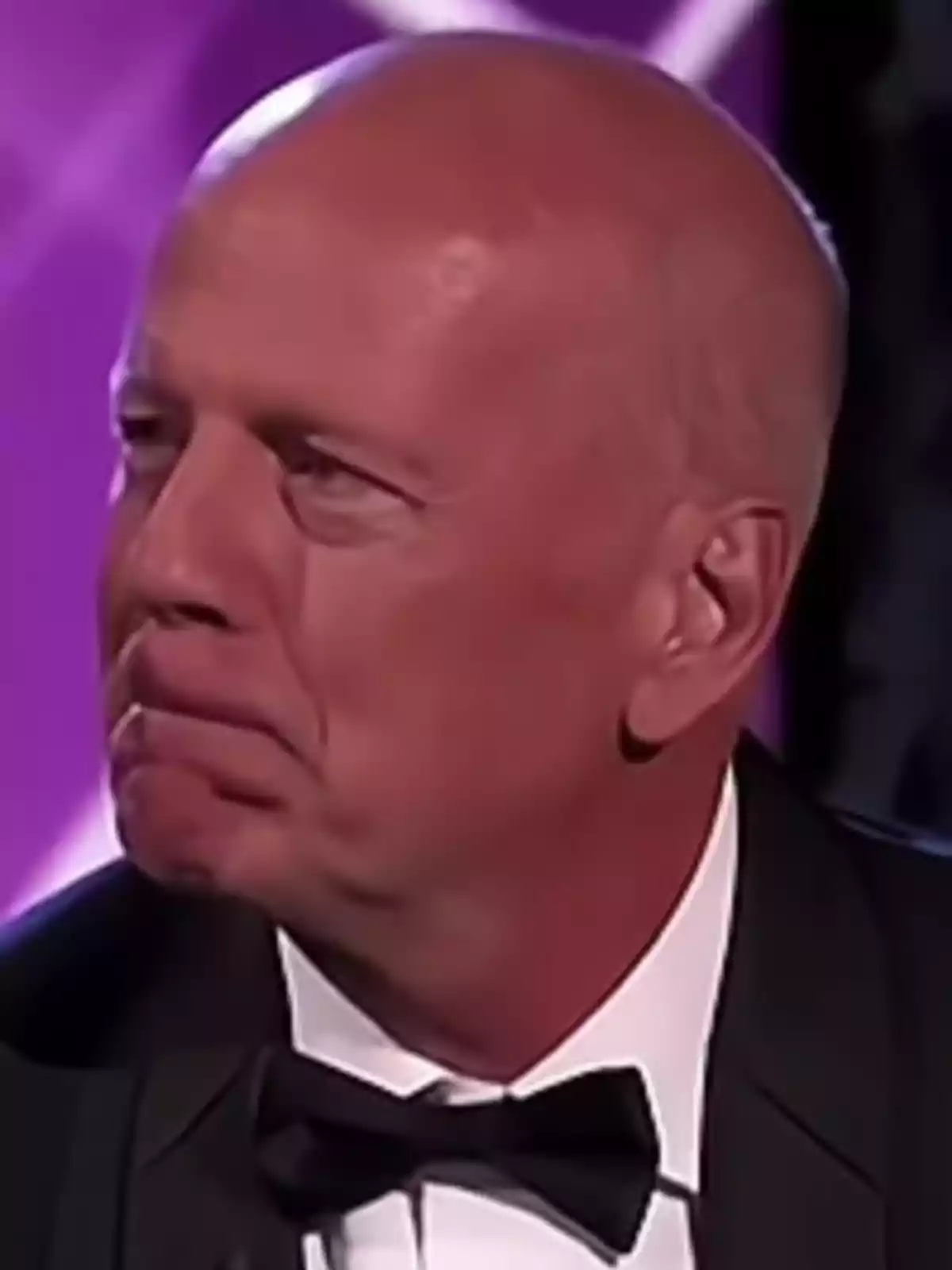 Bruce Willis in a tuxedo and black bow tie at an event with a purple background. Bruce Willis in a tuxedo and black bow tie at an event with a purple background.