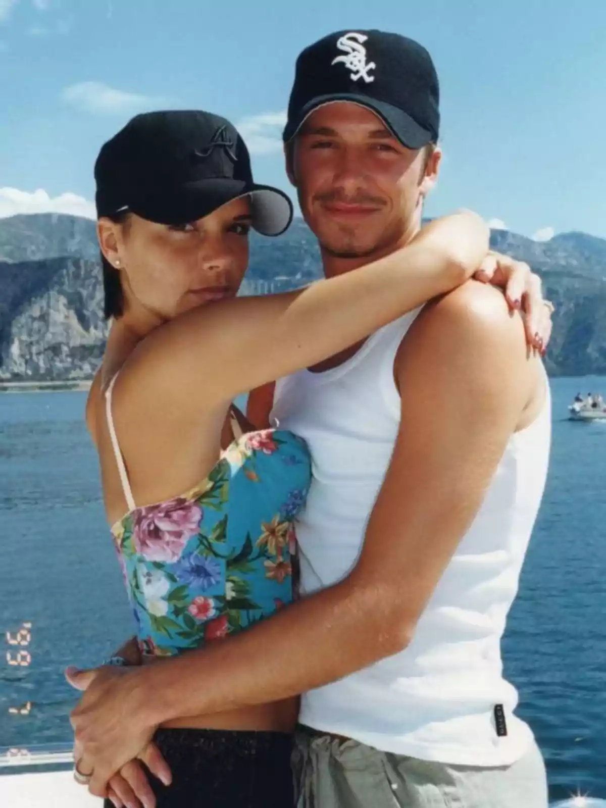 Victoria and David Beckham embrace on a boat with a mountainous landscape and the sea in the background, both wearing black caps and summer clothing. Victoria and David Beckham embrace on a boat with a mountainous landscape and the sea in the background, both wearing black caps and summer clothing.