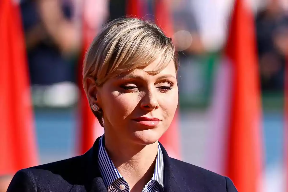 Charlene with short blonde hair, closed eyes and serene expression, dressed in a striped shirt and a dark jacket, with a background of out-of-focus red flags.