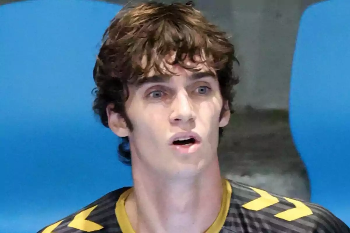Pablo Urdangarin with a surprised expression and curly hair, wearing a black sports jersey with yellow details, sitting in front of a blue background. Pablo Urdangarin with a surprised expression and curly hair, wearing a black sports jersey with yellow details, sitting in front of a blue background.