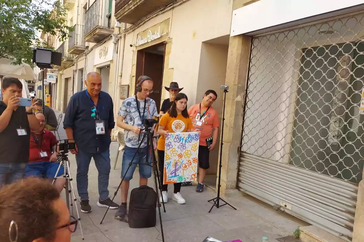 Un grup de persones amb equip d'enregistrament i càmeres es troben en un carrer davant d'una clínica dental, una sosté un cartell colorit amb dibuixos de flors.