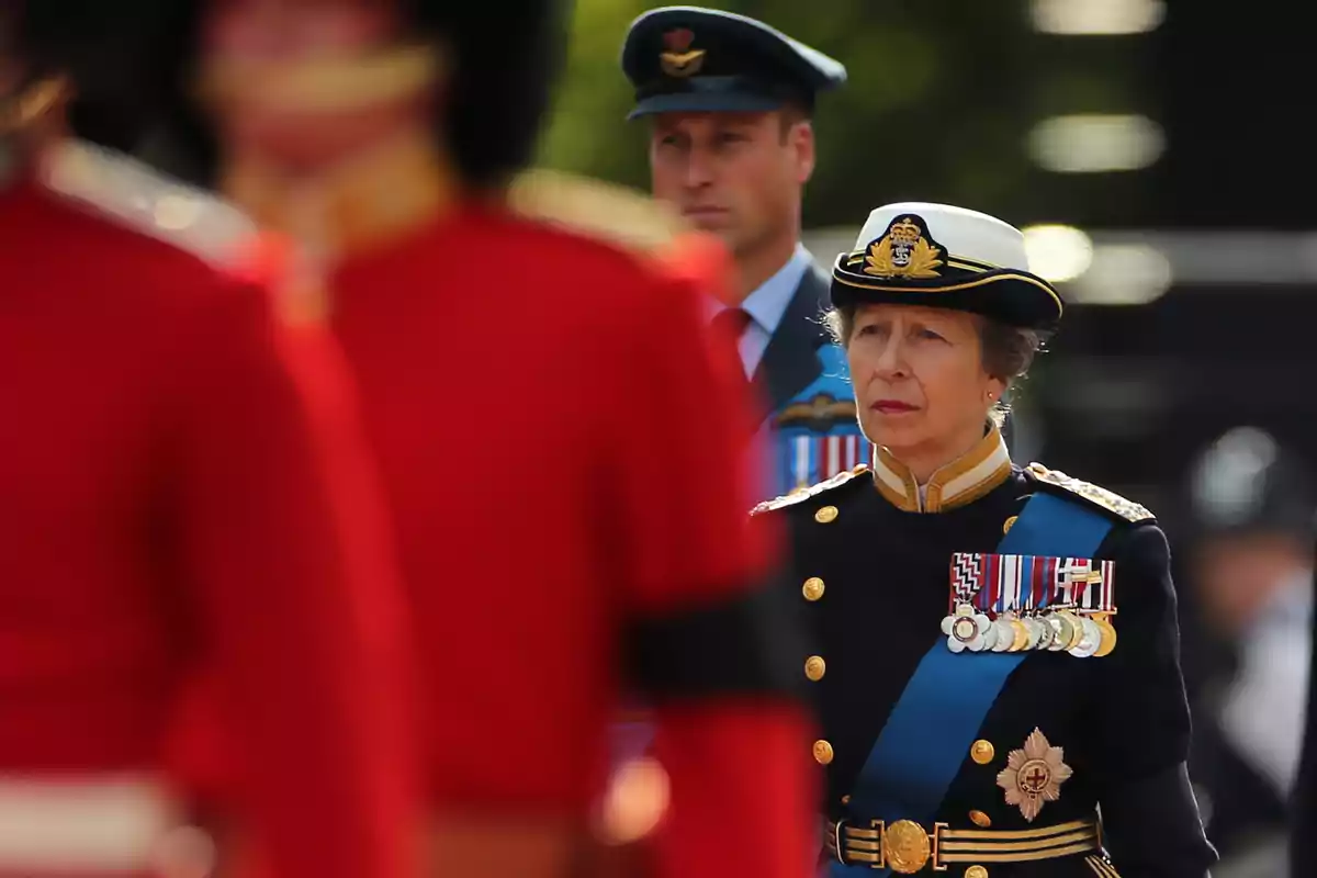 Princess Anne in military uniform and medals walks alongside Prince William in an air force uniform, while soldiers in red uniforms are in the foreground. Princess Anne in military uniform and medals walks alongside Prince William in an air force uniform, while soldiers in red uniforms are in the foreground.