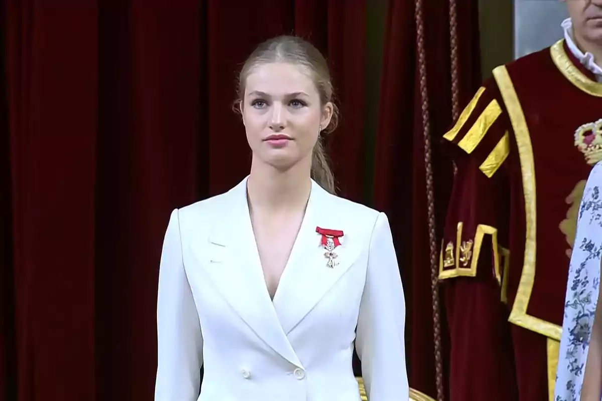 Una mujer con traje blanco y una medalla roja en el pecho, de pie frente a un fondo de cortinas rojas, con una persona vestida con un traje ceremonial detrás de ella.