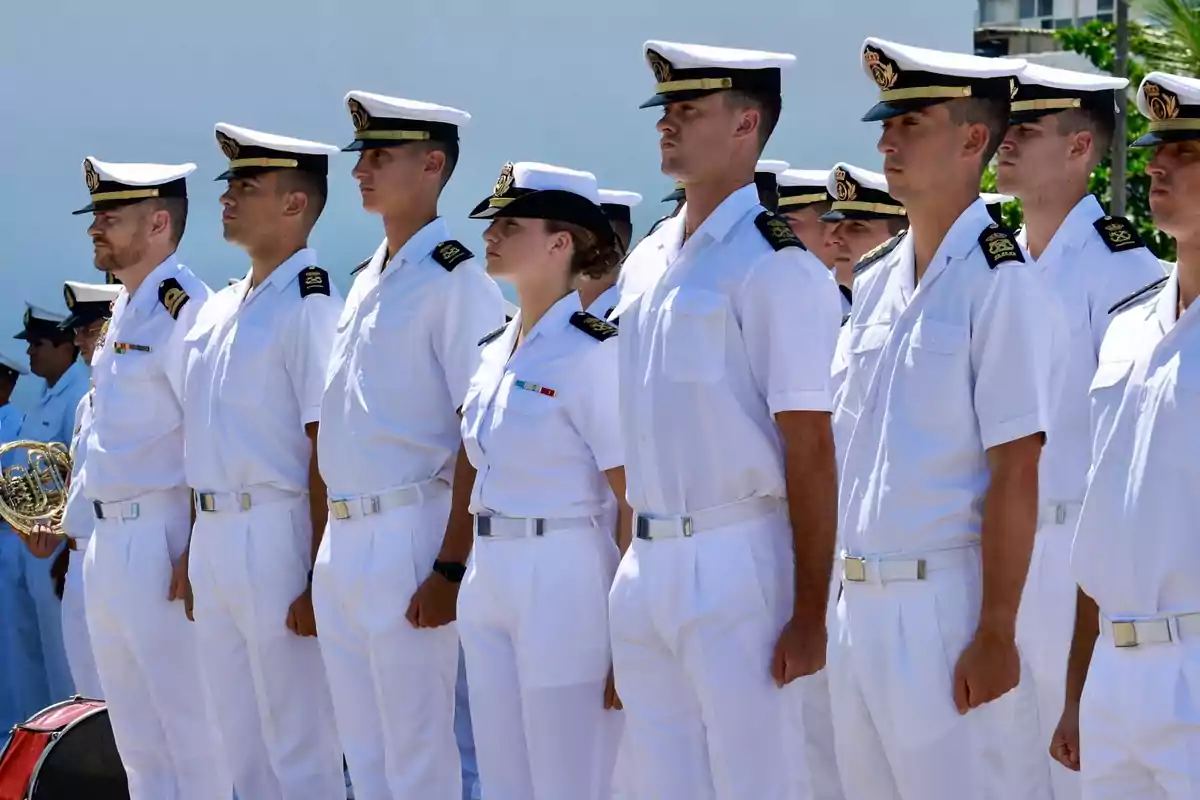 Un grupo de personas vestidas con uniformes blancos de la marina están formados en fila durante una ceremonia al aire libre. Un grupo de personas vestidas con uniformes blancos de la marina están formados en fila durante una ceremonia al aire libre.