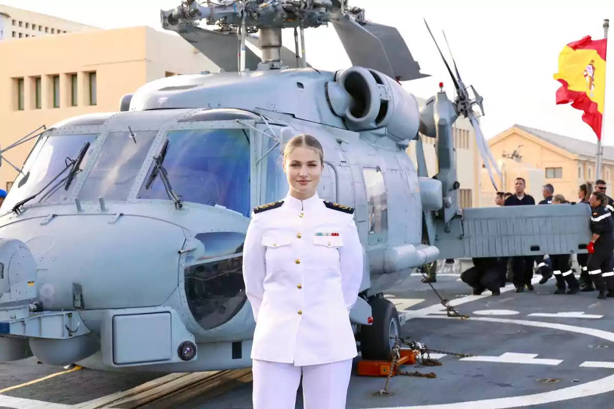 Leonor in a white military dress uniform posing in front of a helicopter at a naval base with the Spanish flag in the background Leonor in a white military dress uniform posing in front of a helicopter at a naval base with the Spanish flag in the background