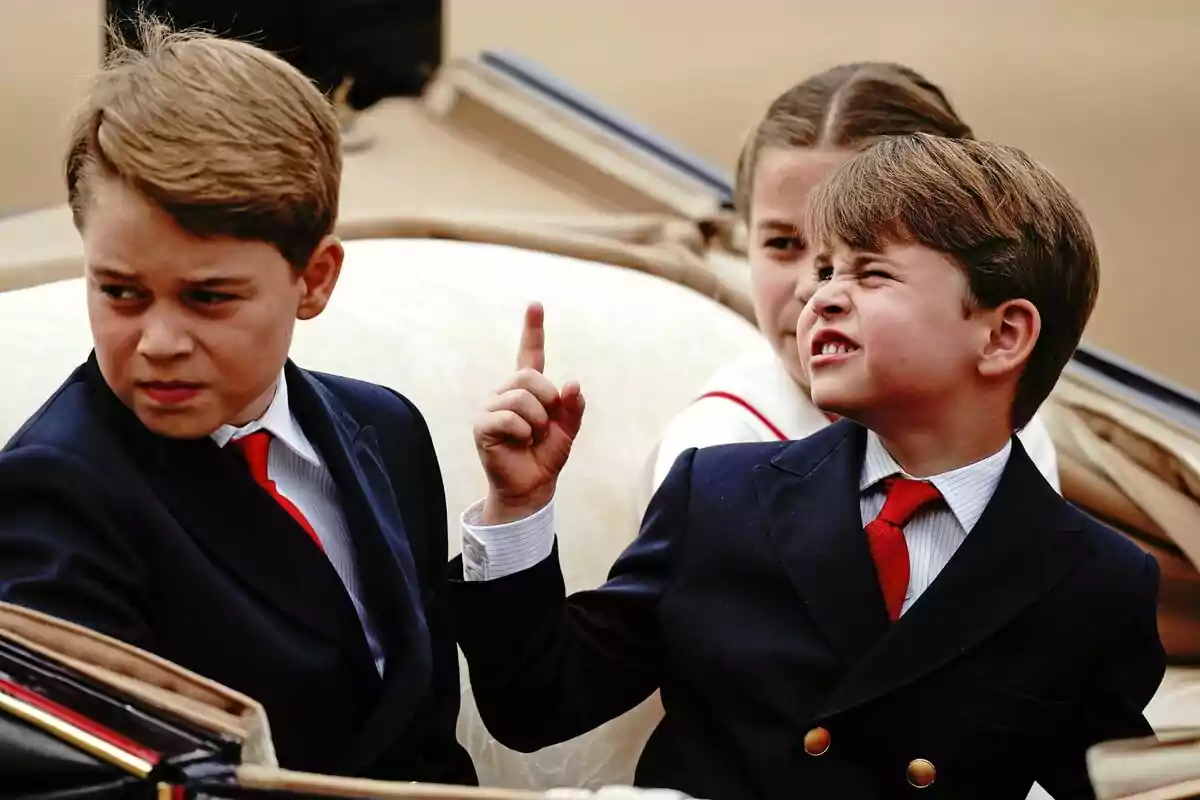 The formally dressed Princes George, Louis and Charlotte are sitting in a carriage, Louis is raising his index finger while grimacing. The formally dressed Princes George, Louis and Charlotte are sitting in a carriage, Louis is raising his index finger while grimacing.