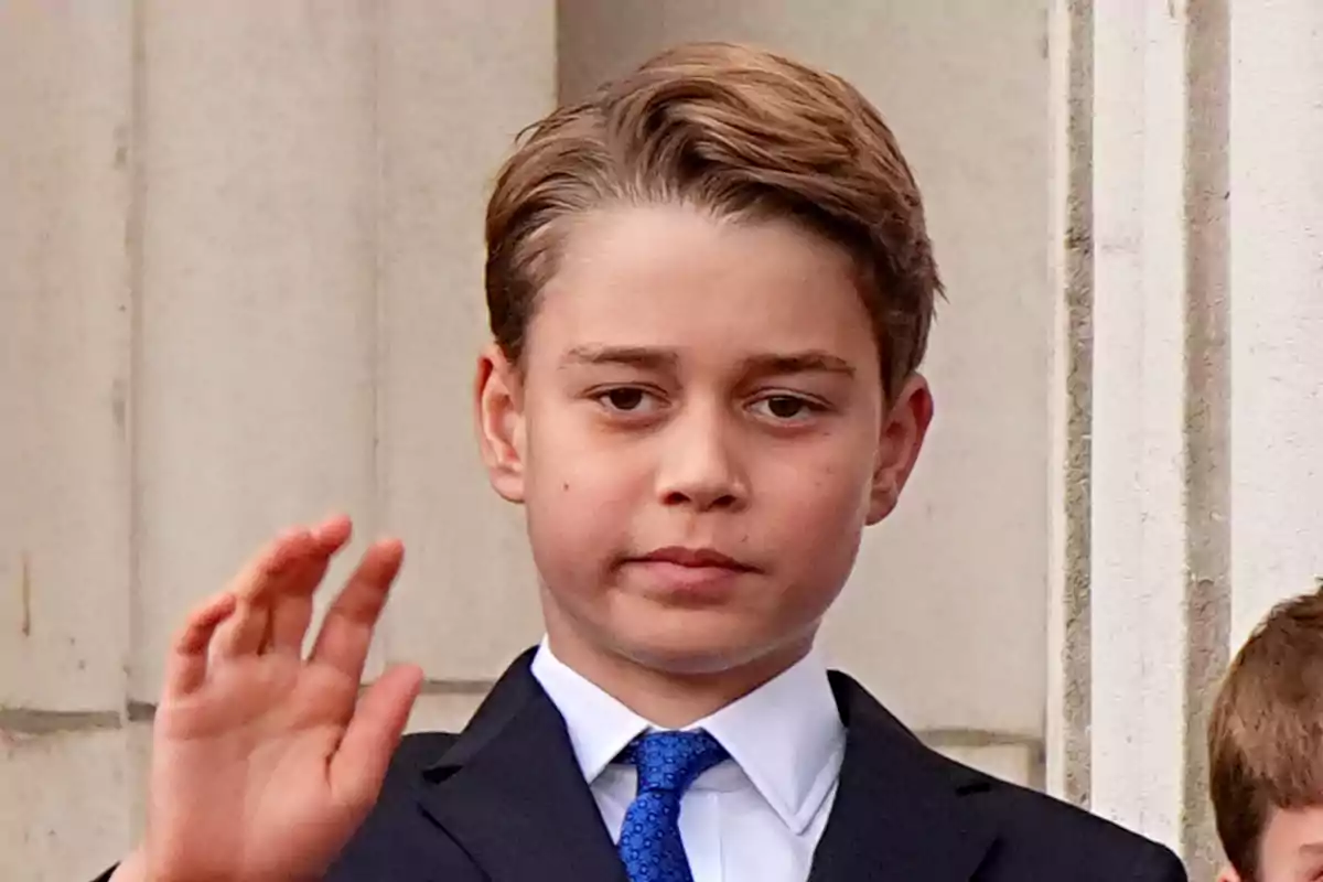 El príncep George amb vestit fosc i corbata blava aixeca la mà davant d'un edifici de pedra. El príncep George amb vestit fosc i corbata blava aixeca la mà davant d'un edifici de pedra.