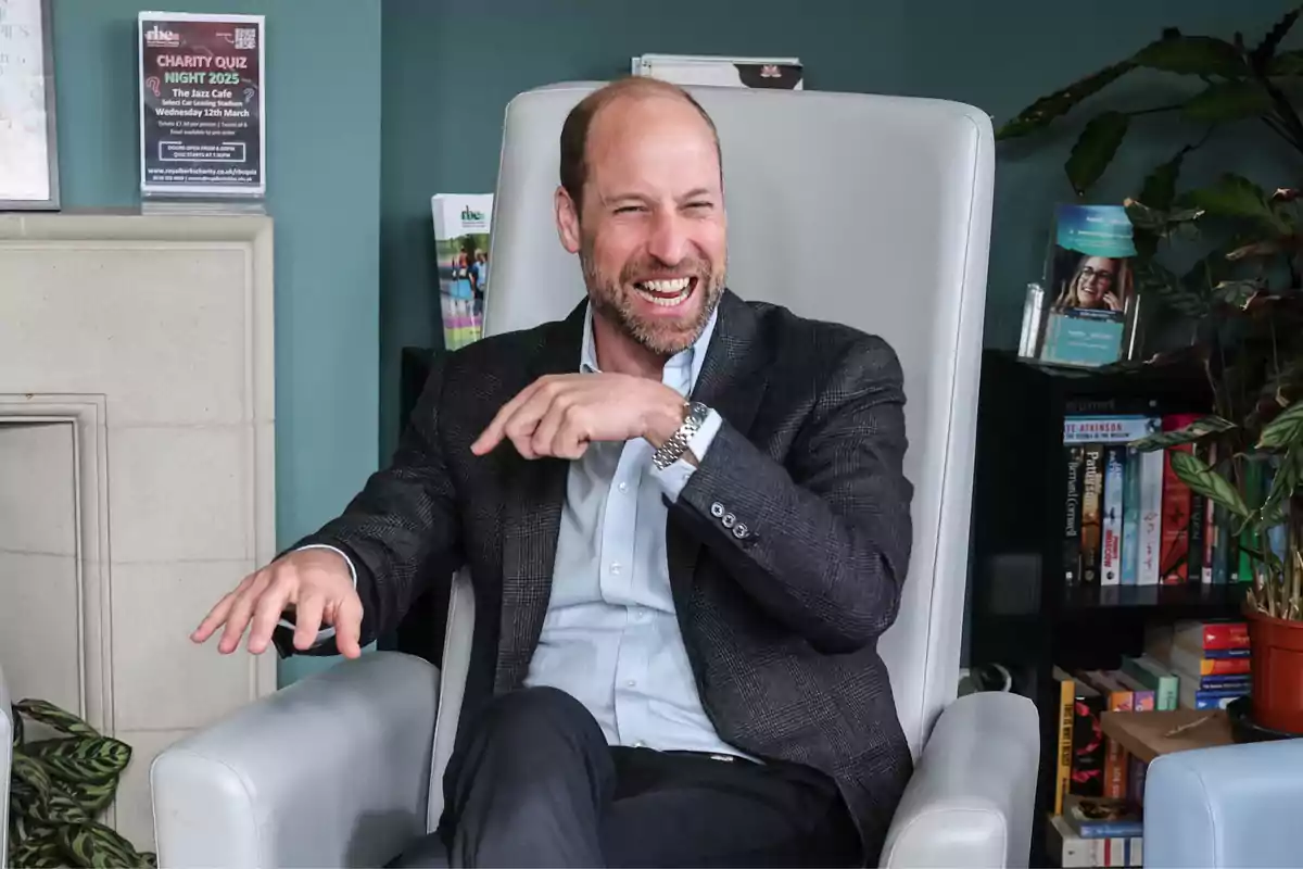 Prince William sitting in a gray chair, smiling and gesturing with his hand, in a setting that appears to be a room with plants and books. Prince William sitting in a gray chair, smiling and gesturing with his hand, in a setting that appears to be a room with plants and books.