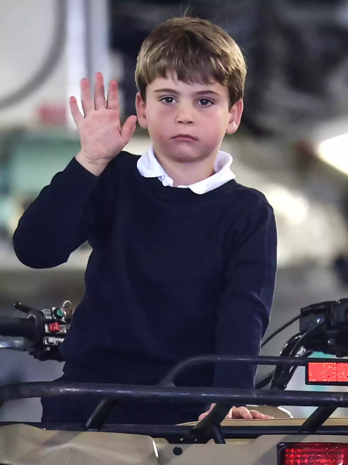 El príncipe Louis con suéter azul oscuro y camisa blanca levanta la mano mientras está de pie sobre un vehículo. El príncipe Louis con suéter azul oscuro y camisa blanca levanta la mano mientras está de pie sobre un vehículo.