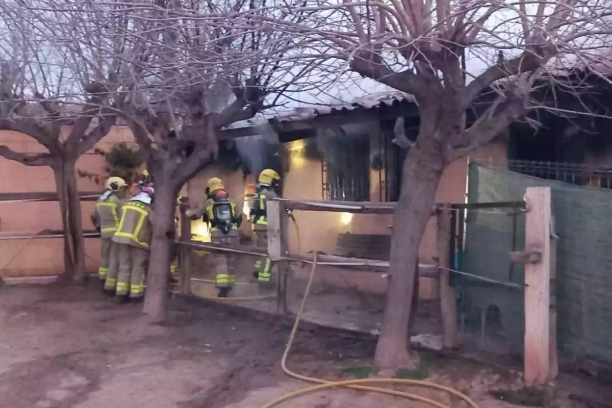 Bombers treballant en un incendi en una casa envoltada d'arbres sense fulles.