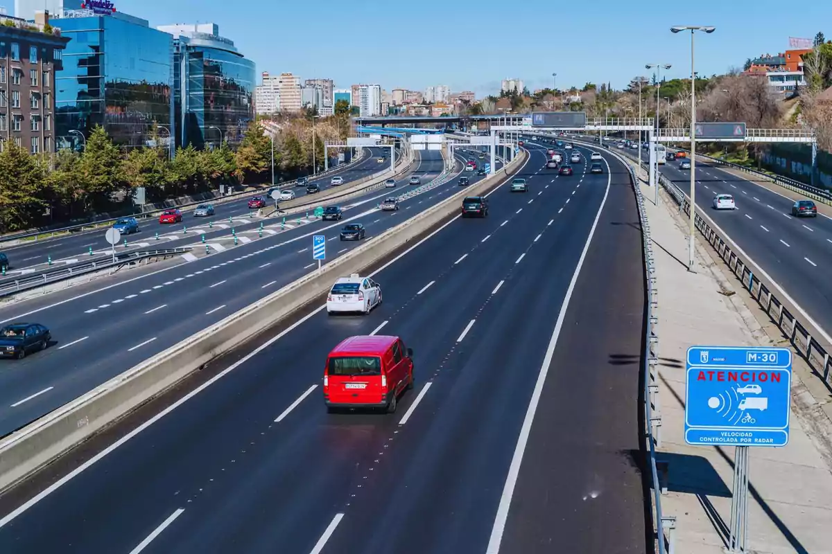 Autopista urbana amb diversos carrils i trànsit moderat, edificis moderns al fons i un senyal de radar de velocitat en primer pla. Autopista urbana amb diversos carrils i trànsit moderat, edificis moderns al fons i un senyal de radar de velocitat en primer pla.
