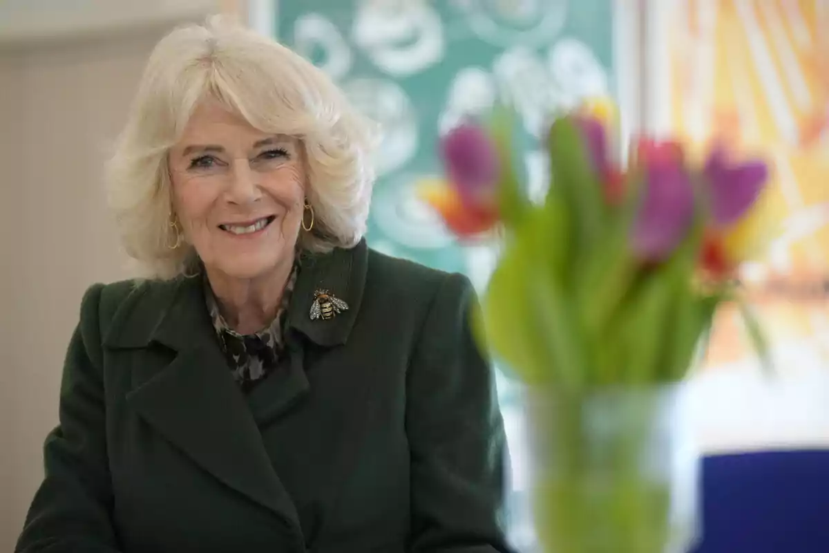 Queen Camila, smiling with blonde hair and a dark green coat with a bee brooch, is sitting in front of a bouquet of colorful tulips. Queen Camila, smiling with blonde hair and a dark green coat with a bee brooch, is sitting in front of a bouquet of colorful tulips.