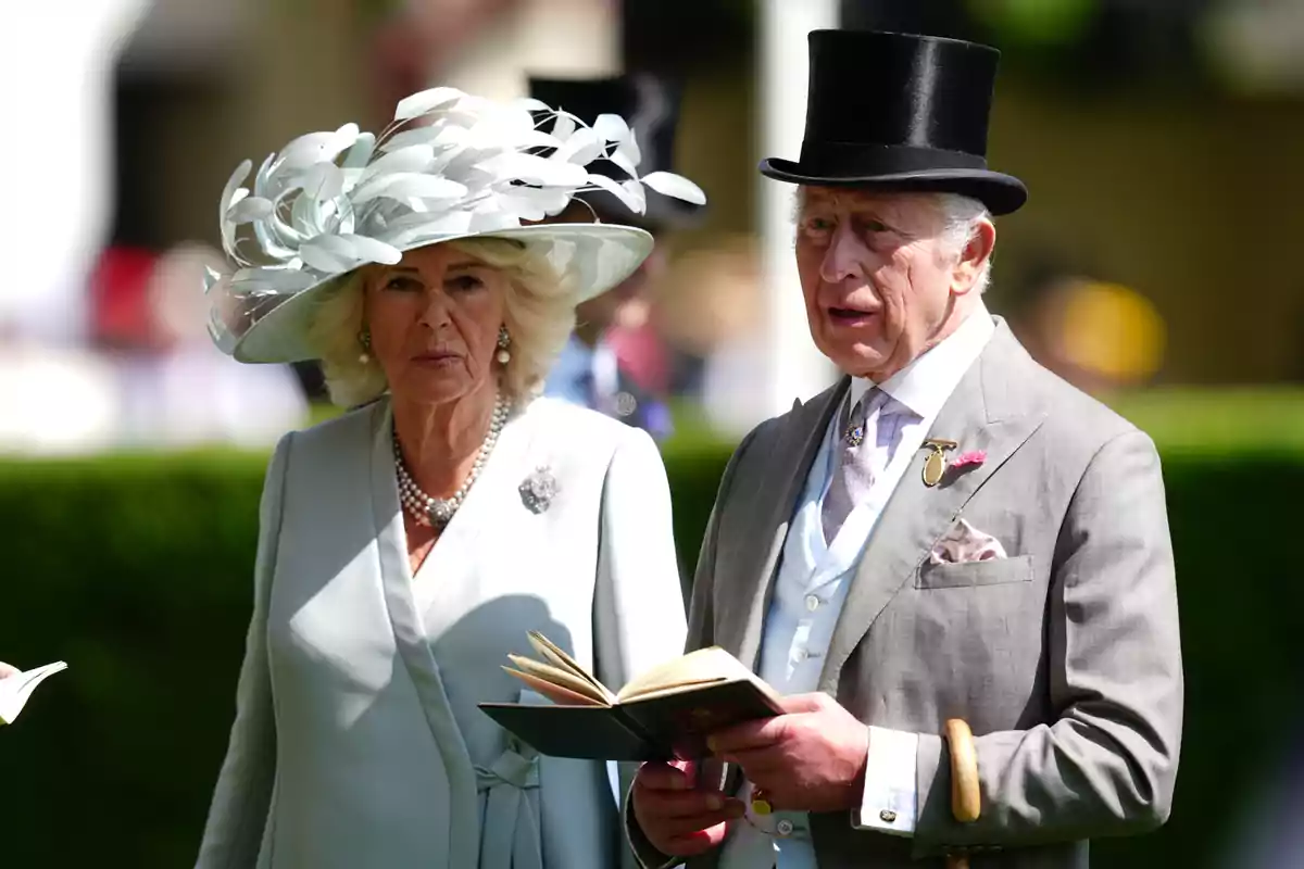 La reina Camila i Carles III elegantment vestits assisteixen a un esdeveniment a l’aire lliure; la dona porta un barret decoratiu blanc i l’home un barret de copa negre i sosté un llibre. La reina Camila i Carles III elegantment vestits assisteixen a un esdeveniment a l’aire lliure; la dona porta un barret decoratiu blanc i l’home un barret de copa negre i sosté un llibre.