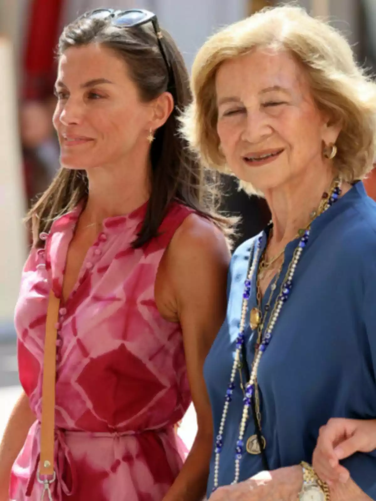 La reina Letícia i la reina Sofia somriuen mentre caminen juntes a l'aire lliure, una porta un vestit rosa i l'altra una brusa blava amb collarets. La reina Letícia i la reina Sofia somriuen mentre caminen juntes a l'aire lliure, una porta un vestit rosa i l'altra una brusa blava amb collarets.