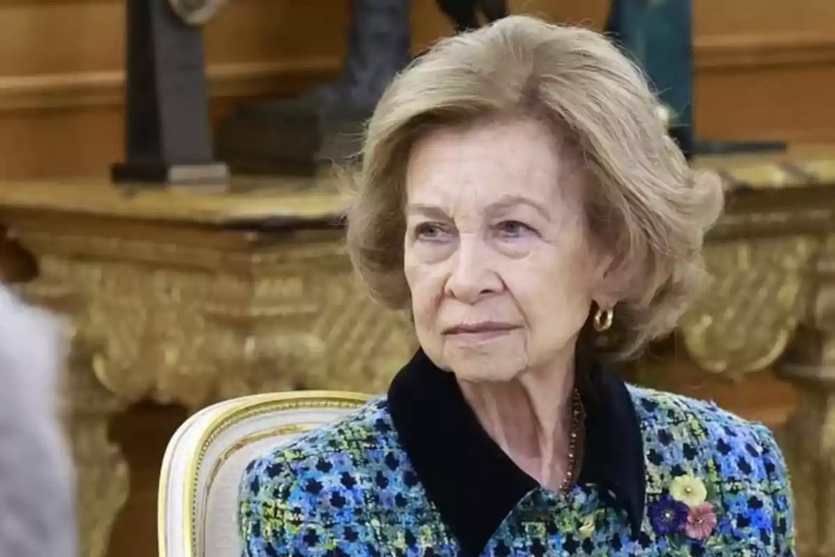 An older woman with blonde hair and elegant hairstyle wears a blue and green colored coat with a flower brooch while sitting on a chair against a background of decorative furniture. An older woman with blonde hair and elegant hairstyle wears a blue and green colored coat with a flower brooch while sitting on a chair against a background of decorative furniture.