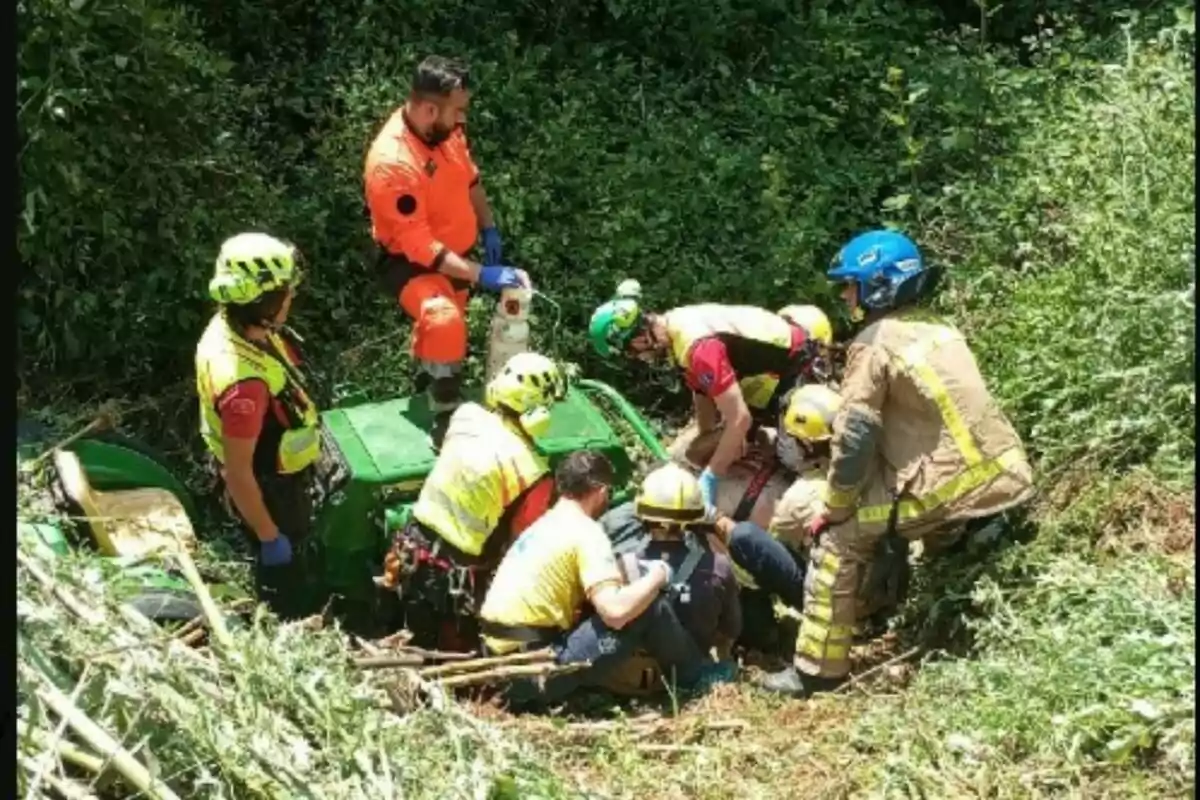 Un grup de rescatadors amb cascos i armilles de seguretat assisteix una persona en una àrea boscosa al costat d'un vehicle verd bolcat.