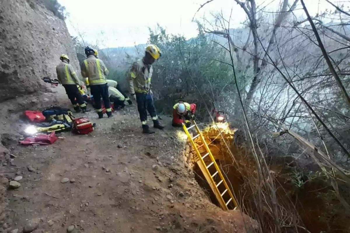 Un grupo de bomberos trabaja en un rescate cerca de un agujero en un terreno montañoso con una escalera amarilla iluminada.