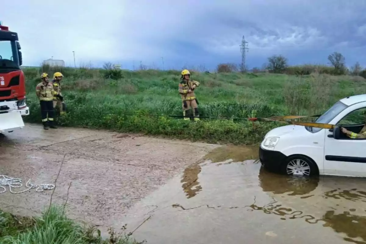 Un grup de bombers amb equip de rescat ajuda un vehicle blanc encallat en un camí inundat, utilitzant un camió de bombers i cordes per remolcar-lo.