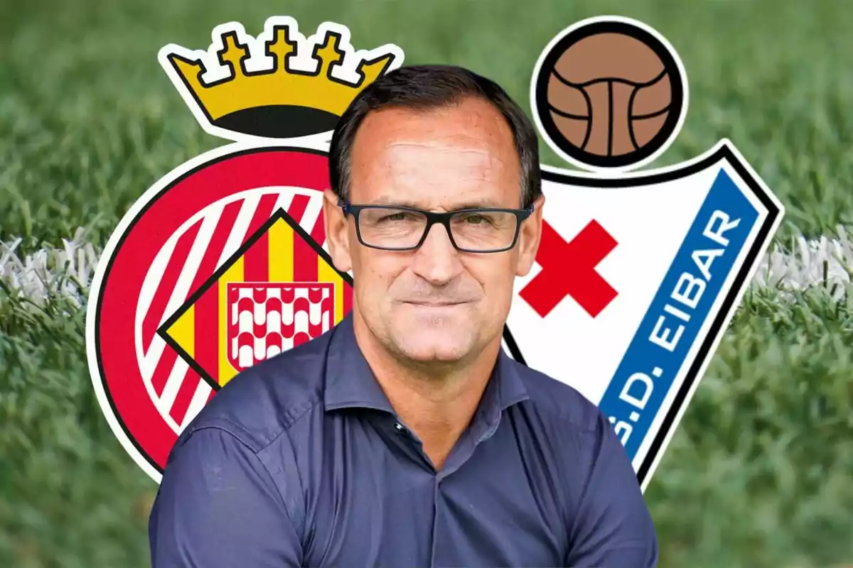 Un hombre con gafas y camisa azul está frente a los escudos de los equipos de fútbol Girona FC y SD Eibar, con un fondo de césped.