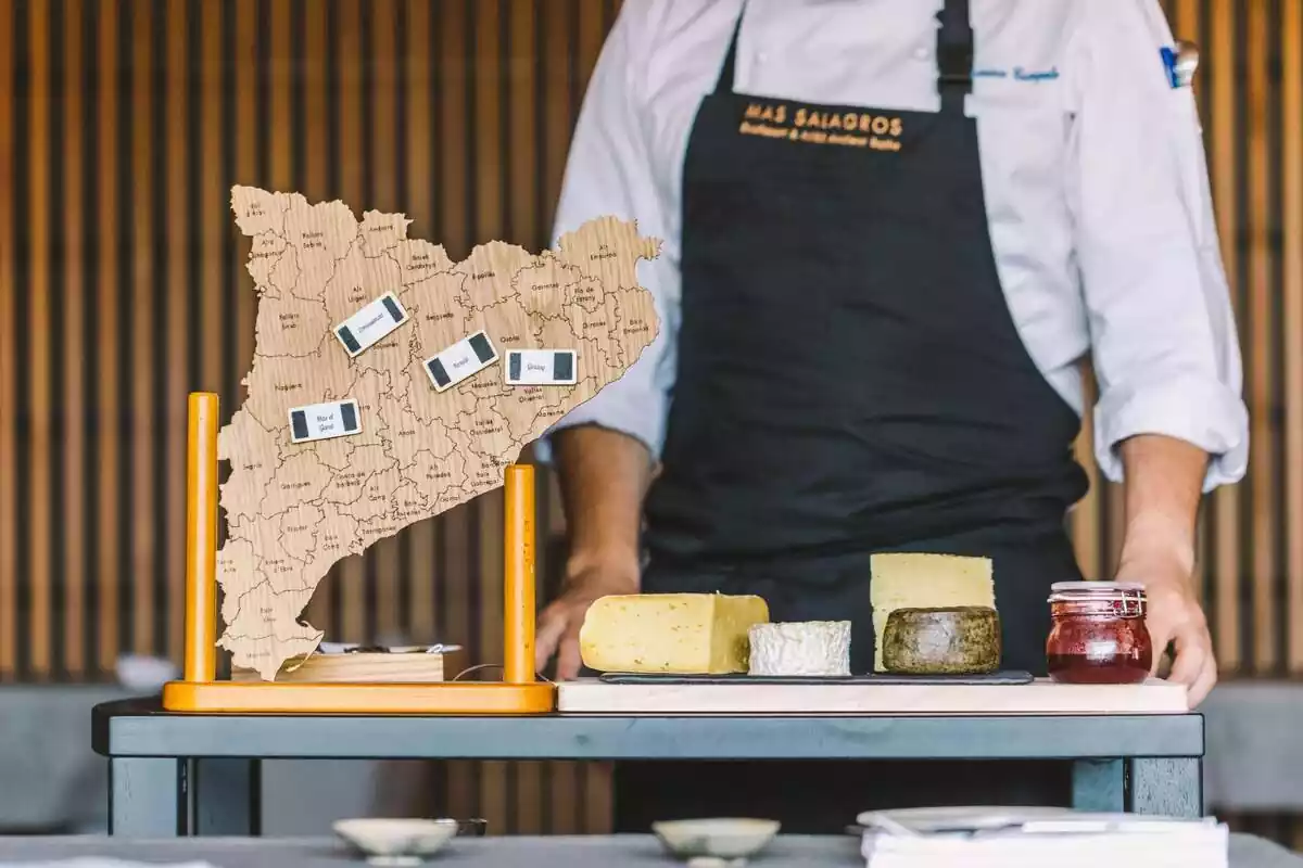 Imatge amb un cuiner darrere una taula de formatges i un mapa de Catalunya