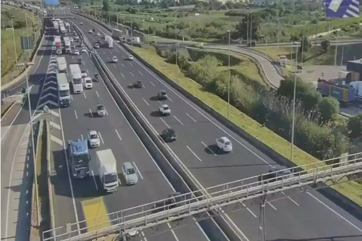 Una autopista amb diversos carrils on circulen automòbils i camions en ambdues direccions, envoltada d'àrees verdes i amb un pas elevat per a vianants.