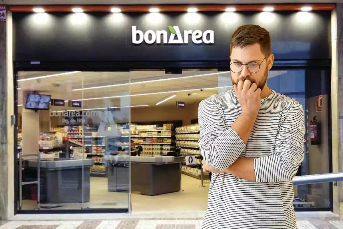 Un hombre con gafas y camiseta de rayas está de pie frente a una tienda bonÀrea, con una expresión pensativa mientras se muerde una uña.