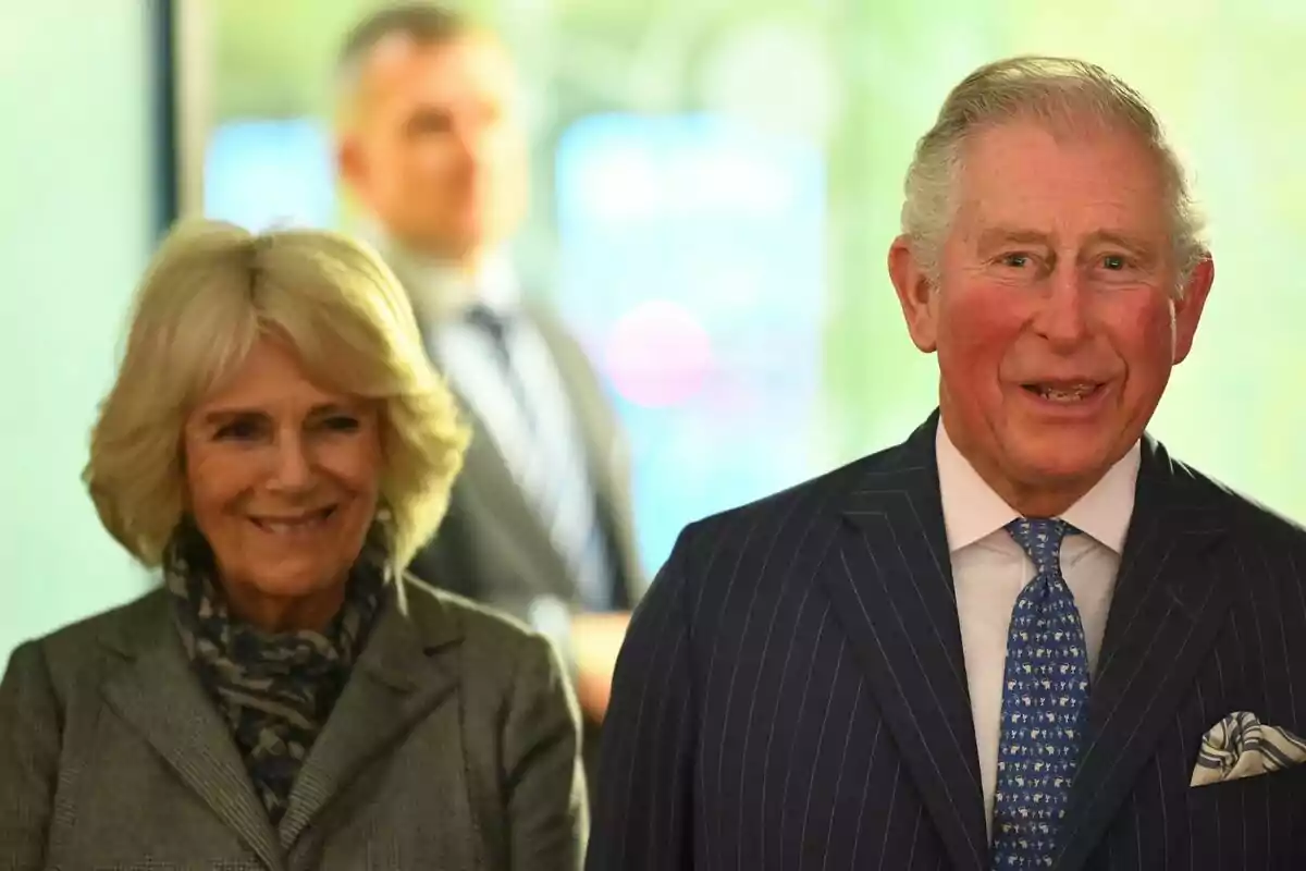 Dues persones grans -que són el rei Carles III i Camilla Parker- somriuen mentre estan dretes, amb un fons desenfocat i una tercera persona al fons. Dues persones grans -que són el rei Carles III i Camilla Parker- somriuen mentre estan dretes, amb un fons desenfocat i una tercera persona al fons.